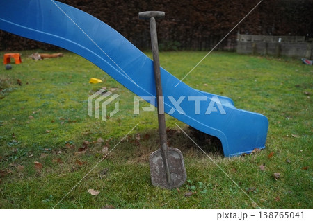 Metal shovel standing in grass beside a blue playground slide on a yard. Outdoor play area scene. Metal shovel standing in grass beside a blue playground slide on a yard. Outdoor play area scene. 138765041