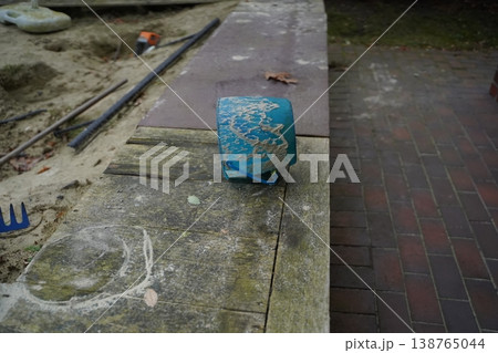 Blue taped cylinder with sand on a weathered ledge beside a brick path and sandy ground; faint chalk marks visible. Outdoor construction scene with small object. 138765044