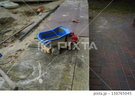 Mud-covered toy dump truck on a weathered wooden platform beside sand and a wet brick walkway. A small child's play vehicle left outdoors after play. Mud-covered toy dump truck on a weathered wooden platform beside sand and a wet brick walkway. A small child's play vehicle left outdoors after play. 138765045