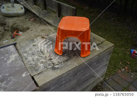 Orange plastic stool on a worn wooden sandbox edge beside sand and yard pavers. A small outdoor seat sits near a play area. Orange plastic stool on a worn wooden sandbox edge beside sand and yard pavers. A small outdoor seat sits near a play area. 138765050