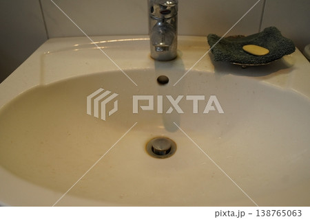 White bathroom sink with chrome faucet, drain, and overflow hole; a green scrub pad and yellow soap sit on the rim. A simple washbasin scene. 138765063