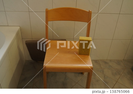 Wooden chair with a yellow sponge on the seat in a tiled bathroom near a bathtub. Everyday cleaning scene. Wooden chair with a yellow sponge on the seat in a tiled bathroom near a bathtub. Everyday cleaning scene. 138765066