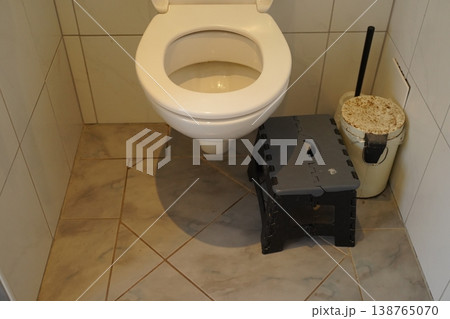 Toilet in a tiled bathroom with a plastic step stool and a covered bucket beside a cleaning brush. Simple restroom scene. 138765070