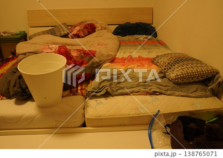 Messy bedroom with low bed and rumpled blankets, pillows, and stained fabric; a white bucket sits on a mattress with scattered clothes and bags. The room looks untidy. Messy bedroom with low bed and rumpled blankets, pillows, and stained fabric; a white bucket sits on a mattress with scattered clothes and bags. The room looks untidy. 138765071