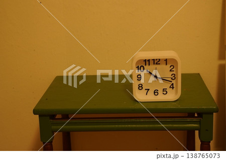 White analog alarm clock on a green wooden table against a beige wall, showing numbers "1 2 3 4 5 6 7 8 9 10 11 12". A simple indoor still life. 138765073