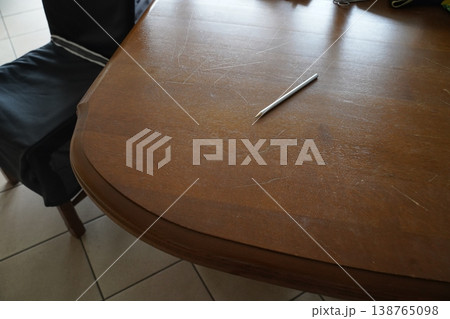 Scratched wooden table with a silver pencil lying on it beside a dark chair on tiled floor in daylight. An empty tabletop scene. Scratched wooden table with a silver pencil lying on it beside a dark chair on tiled floor in daylight. An empty tabletop scene. 138765098
