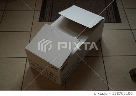 Open white cardboard box with flaps on a tiled floor; small printed text on side is unreadable. An empty package sits on the floor indoors. 138765100