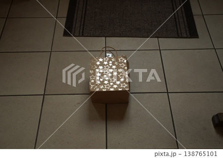 Gold gift box with star pattern and a loop handle on tiled floor near a dark mat. A present awaits on the floor. 138765101