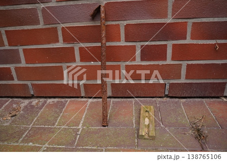 Rusty metal rod leaning against brick wall above tiled pavement with a small ground fixture and dry twigs 138765106