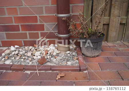 Brick wall with drainpipe, gravel bed, paving stones, and a flowerpot with dried plants beside a wooden fence 138765112