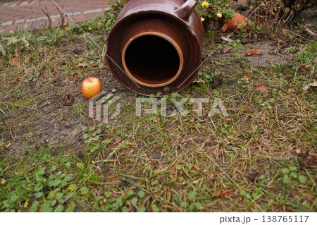 Brown ceramic jar lying on its side on patchy grass and soil, with a red apple nearby 138765117