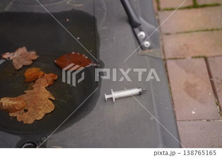 Syringe on a dark surface near a puddle with fallen leaves, beside a paved walkway 138765165