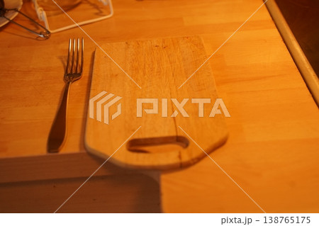 Wooden cutting board on a table with a fork beside it under warm light; blurred kitchen items in background Wooden cutting board on a table with a fork beside it under warm light; blurred kitchen items in background 138765175