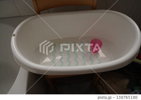 White baby bathtub on a stand with shallow water, blue wavy anti-slip lines, and a pink cup inside in a tiled bathroom 138765180