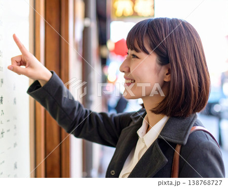 繁華街で食べ歩きを楽しむ若い女性。メニュー看板を指差して微笑む。 繁華街で食べ歩きを楽しむ若い女性。メニュー看板を指差して微笑む。 138768727