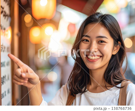 繁華街で食べ歩きを楽しむ若い女性。メニュー看板を指差して微笑む。 繁華街で食べ歩きを楽しむ若い女性。メニュー看板を指差して微笑む。 138768736