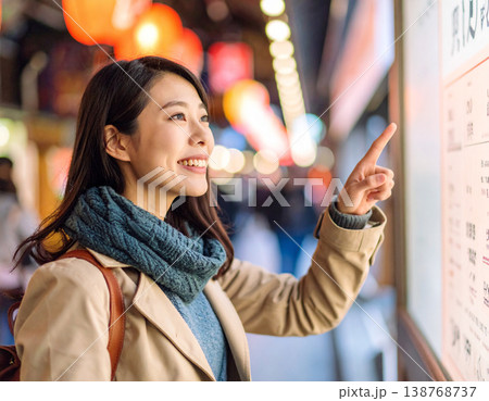 繁華街で食べ歩きを楽しむ若い女性。メニュー看板を指差して微笑む。 繁華街で食べ歩きを楽しむ若い女性。メニュー看板を指差して微笑む。 138768737