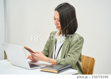 オフィスのデスクでノートPCを開きスマートフォンを操作する女性社員 138778399