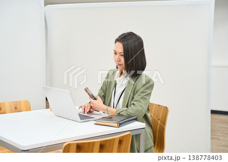 オフィスのデスクでノートPCを開きスマートフォンを操作する女性社員 138778403