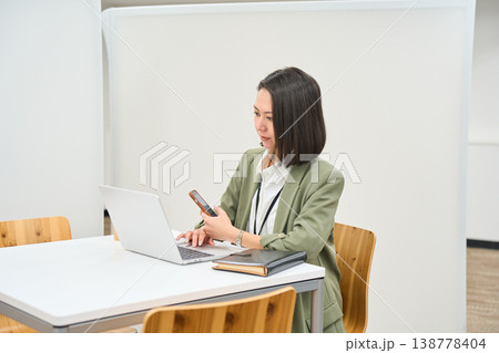 オフィスのデスクでノートPCを開きスマートフォンを操作する女性社員 138778404
