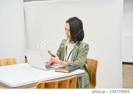 オフィスのデスクでノートPCを開きスマートフォンを操作する女性社員 オフィスのデスクでノートPCを開きスマートフォンを操作する女性社員 138778405