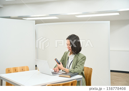 オフィスのデスクでノートPCを開きスマートフォンを操作する女性社員 138778408