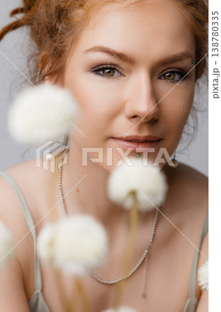 Young Caucasian redhead woman touching face among dried white flowers on moss, white wall Young Caucasian redhead woman touching face among dried white flowers on moss, white wall 138780335