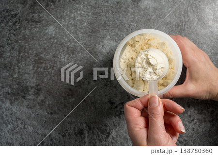 Female hands holding a scoop of protein whey powder, white plastic tube, on dark background, copy space 138780360