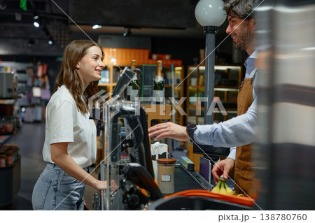 A man cashier wearing an apron is engaging in conversation with a woman in a store at cash register A man cashier wearing an apron is engaging in conversation with a woman in a store at cash register 138780760