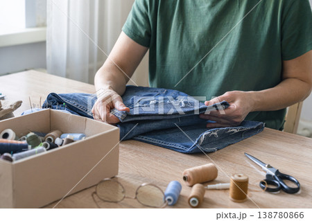 Person hands holding and mending ripped denim jeans on a wooden table, embracing eco-friendly repair and upcycling, for sustainable fashion. Reusing things Person hands holding and mending ripped denim jeans on a wooden table, embracing eco-friendly repair and upcycling, for sustainable fashion. Reusing things 138780866
