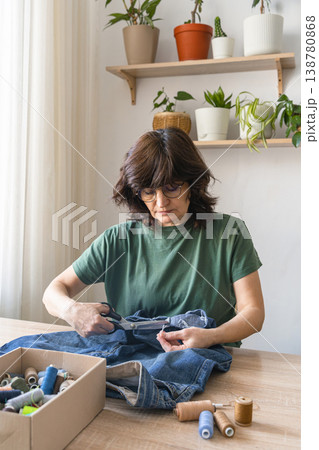 An adult woman cutting thread from old jeans for reuse, promoting sustainable fashion and circular economy principles. Person upcycling denim jeans with scissors, repairing clothing An adult woman cutting thread from old jeans for reuse, promoting sustainable fashion and circular economy principles. Person upcycling denim jeans with scissors, repairing clothing 138780868