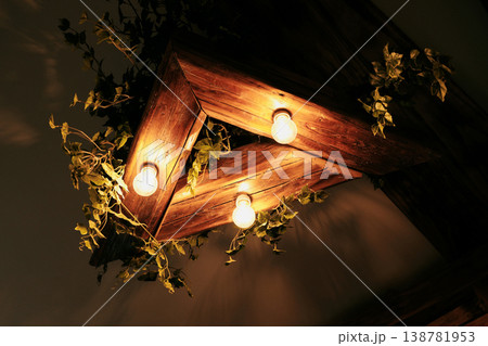 Decorative wooden ceiling light fixture with warm bulbs and green plants. Concept of interior design, ambiance lighting and cozy atmosphere Decorative wooden ceiling light fixture with warm bulbs and green plants. Concept of interior design, ambiance lighting and cozy atmosphere 138781953