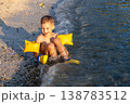 Happy child sits sandy beach shoreline small waves splash wearing bright inflatable armbands under warm golden sunlight. Playful summer vacation scene joy freedom leisure carefree childhood by sea 138783512