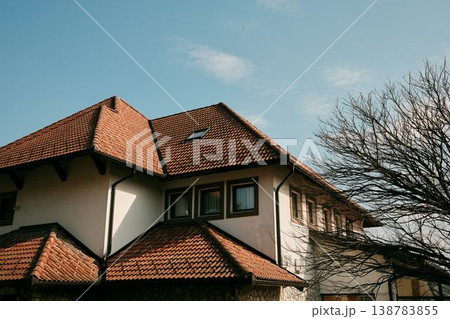 Residential building with traditional red clay tile roof and bare tree branches under blue sky in Balkans, Bosnia. Concept of suburban architecture, housing and peaceful neighborhood life Residential building with traditional red clay tile roof and bare tree branches under blue sky in Balkans, Bosnia. Concept of suburban architecture, housing and peaceful neighborhood life 138783855