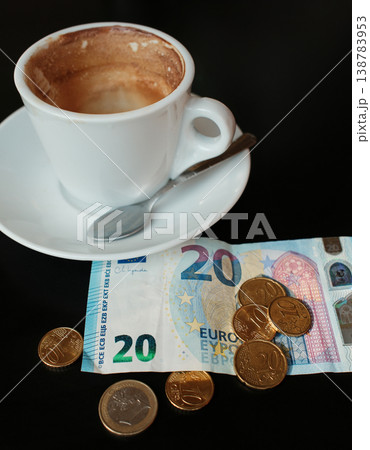 Partially drunk cappuccino in white cup with euro banknote and coins on dark table. Concept of cafe payment, coffee price and everyday spending in European economy Partially drunk cappuccino in white cup with euro banknote and coins on dark table. Concept of cafe payment, coffee price and everyday spending in European economy 138783953