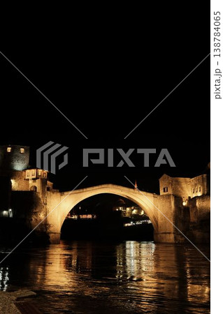 Night view of the Old Bridge in Mostar with illuminated stone arch and medieval towers reflecting in the Neretva River under a dark sky 138784065