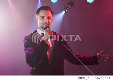 Charismatic showman host singing with microphone on stage under vivid lights Charismatic showman host singing with microphone on stage under vivid lights 138784673