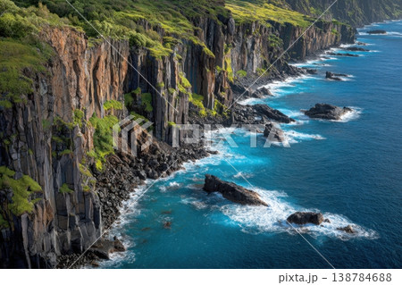 Coastal scenery shows cliffs rising above blue ocean waves. Rocks are visible along the shoreline with green plants on the cliffs 138784688