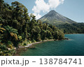 A tall volcano rises next to a clear blue lake surrounded by green trees under a bright sky with some clouds 138784741