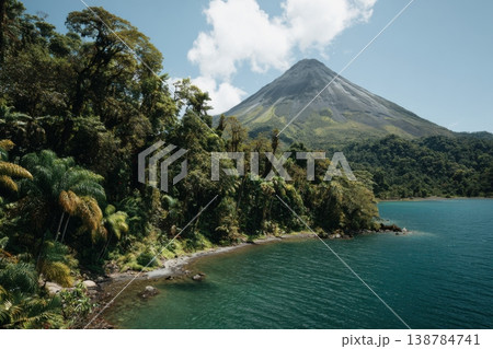 A tall volcano rises next to a clear blue lake surrounded by green trees under a bright sky with some clouds 138784741