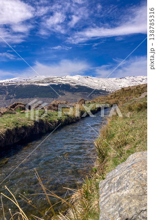 A clear spring stream flows through alpine landscape with snow-capped peaks of Sierra Nevada Mountains lush valley meadow in Spain A clear spring stream flows through alpine landscape with snow-capped peaks of Sierra Nevada Mountains lush valley meadow in Spain 138785316
