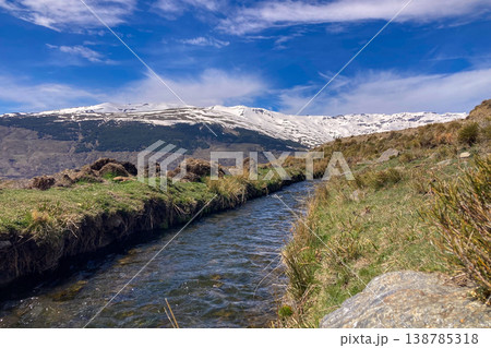 A clear spring stream flows through alpine landscape with snow-capped peaks of Sierra Nevada Mountains lush valley meadow in Spain A clear spring stream flows through alpine landscape with snow-capped peaks of Sierra Nevada Mountains lush valley meadow in Spain 138785318