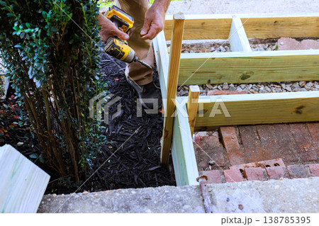Man uses drill to secure wood steps frame near bush in garden area Man uses drill to secure wood steps frame near bush in garden area 138785395