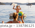 Happy child plays calm sea water inflatable ring mother throws colorful ball warm summer vacation. Bright warm sunlight waves yachts create joyful family atmosphere filled laughter carefree moments 138786125