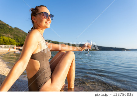Smiling woman in sunglasses relaxes near calm water holding glass during warm sunset. Natural beauty, summer vacation mood, peaceful landscape freedom leisure serene travel atmosphere by quiet shore 138786838