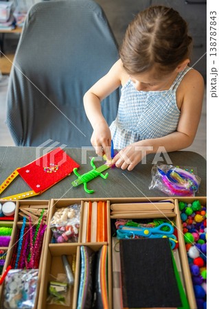 Little girl gluing colorful feathers to handmade craft toy at table with creative supplies. Kids creativity fine motor skills development and learning through play concept. Little girl gluing colorful feathers to handmade craft toy at table with creative supplies. Kids creativity fine motor skills development and learning through play concept. 138787843