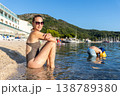 Smiling young adult woman relax bikini sunny beach hold glass wine child play water inflatable arm. Summer vacation lifestyle family carefree leisure mediterranean coast green mountains background 138789380