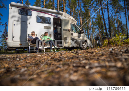 Two adults relaxing by camper van in sunny forest campsite. Two adults relaxing by camper van in sunny forest campsite. 138789633