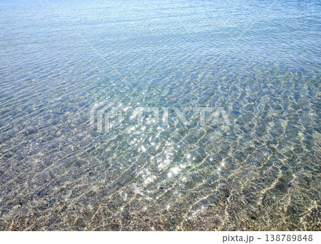 Clean transparent water on shallow sandy seashore with shells with shells on sunny summer day. Sun glare on surface of water. Small waves on sea coast. Nature backdrop. Natural background 138789848