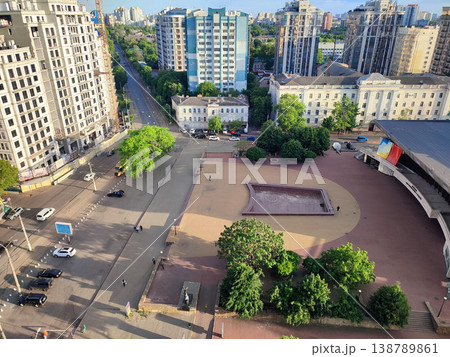 City street view from above. Large multi-story buildings, a square, green trees, an asphalt road for cars, moving cars and walking people on a sunny summer evening City street view from above. Large multi-story buildings, a square, green trees, an asphalt road for cars, moving cars and walking people on a sunny summer evening 138789861